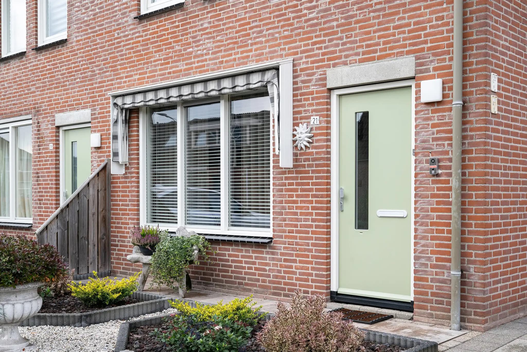 Zachtgroene voordeur naast een raam met markies en een verzorgde voortuin